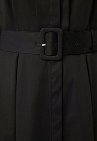 Schwarzes Kleid mit einer taillierten Taille und einem breiten Gürtel mit einer quadratischen Schnalle. Der Stoff ist glatt und hat subtile Falten entlang der Vorderseite.