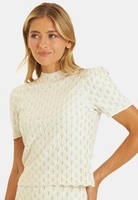 Biały, dziergany top z teksturowanym wzorem nachodzących na siebie diamentów, krótki rękaw i wysoka szyja. Lekki materiał.