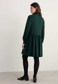 Zielona, długa sukienka z długim rękawem, zebranym w talii, zwiewnym dołem i wysoką szyją. Noszona z czarnymi rajstopami i butami na obcasie, gładka tekstura materiału.