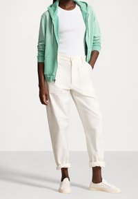 Mátově zelená mikina na zip, bílý přiléhavý top, krémové zúžené kalhoty s ohrnutými manžetami a bílé tenisky.