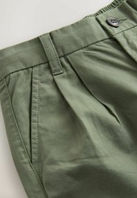 Gros plan de la ceinture d’un pantalon vert avec passants de ceinture, poche latérale, plis et fermeture boutonnée sur fond clair.