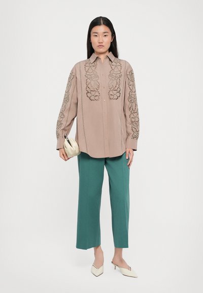 Hellbeige Button-Up-Hemd mit floralem Stickmuster, kombiniert mit türkisfarbenen, weit geschnittenen Hosen und einer cremefarbenen Clutch. Klassische spitze Absatzschuhe.