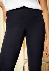 Nő magas derekú, fekete, testhezálló nadrágot és fehér felsőt visel, egyik kezét a zsebében tartja, az elmosódott háttérben fém szék látható.