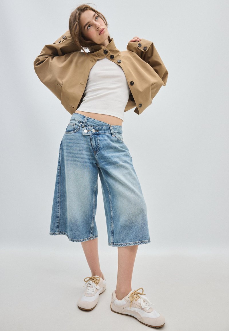 Jeune femme s'étirant dans une veste beige oversize, un crop top blanc, un short en jean large et des baskets blanches sur un fond clair uni.