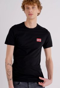Melna kokvilnas T-krekls ar īsām piedurknēm un apaļu kakla izgriezumu, ar mazu sarkanu Levi's logu uz kreisā krūšu.