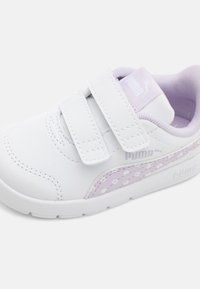 Baltas sporta apavi ar purpura akcentiem, Velcro siksnām, gludu tekstūru, noapaļotu purngala daļu un punktveida rakstu sānos. Redzams PUMA logo.