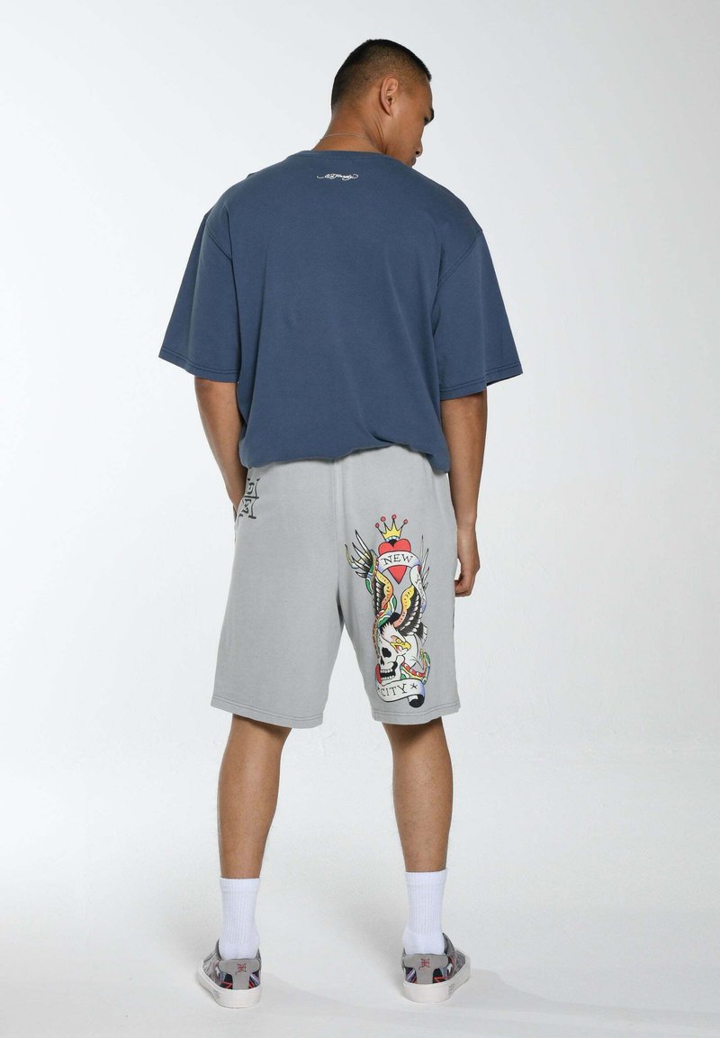T-shirt in cotone blu navy rilassato abbinato a pantaloni corti grigi con una grafica colorata in stile tatuaggio di un teschio e ali sul retro.
