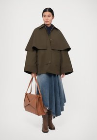 Olive green oversized jacket with a cape silhouette, paired with a flared blue denim skirt. Brown leather handbag and knee-high brown boots.
