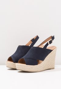 Marinblå kilklackar med en mjuk textiloversida, öppen tå-design och flätad juteplattform för textur och kontrast. Justerbart ankelband.
