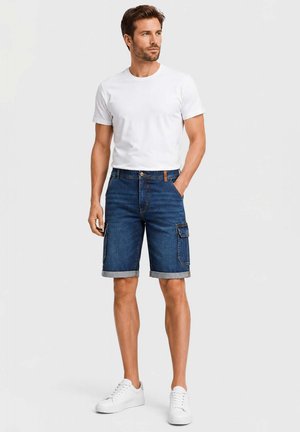 Man standing with one hand in pocket, wearing white t-shirt, rolled-up blue denim cargo shorts, and white sneakers.