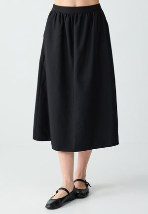 Femme portant une jupe midi noire à taille élastique et des ballerines Mary Jane noires, sur un fond clair uni.