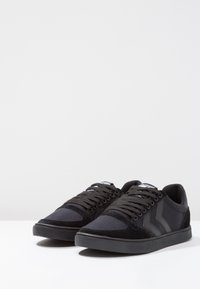 Tenis pretos com uma combinação de materiais de camurça e tecido, apresentando atacadores pretos, uma sola de borracha e um padrão de chevron distinto na lateral.