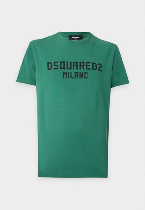 Homme portant un t-shirt vert Dsquared2 Milano, un short en jean jusqu'aux genoux avec chaîne, des chaussettes blanches et des baskets noires, debout avec une main dans la poche.