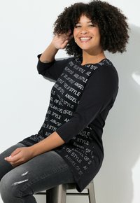 Schwarz-langärmliges Oberteil mit weißem Textmuster "ANGEL OF STYLE", kombiniert mit schwarzen Ärmeln; getragen mit dunklen Jeans. Modell sitzt auf einem Hocker.