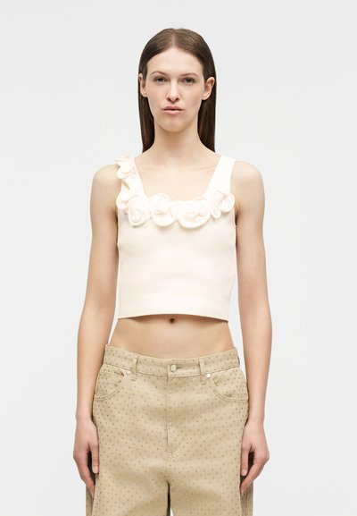 Νεαρή γυναίκα με μακριά καστανά μαλλιά φοράει ένα λευκό cropped αμάνικο τοπ με βολάν και λουλουδένιες λεπτομέρειες και μπεζ ψηλόμεσο παντελόνι με μικρά σχέδια.