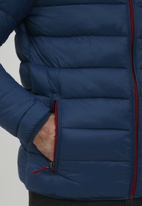 Jūros jėgų pūkuota striukė su siuvinėtu tekstūru ir kontrastinga raudona užtrauktuko detale. Apima šonines kišenes ir aukštą apykaklę papildomam šilumui.