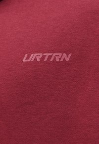 Közeli kép egy piros anyagról, amelynek közepén az "URTRN" szó világosabb piros betűkkel van nyomtatva.