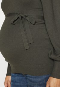 Maglia lavorata a maglia verde scuro con laccio in vita, aderente all'addome. Presenta una texture a costine e maniche lunghe con spalle leggermente a sbuffo.