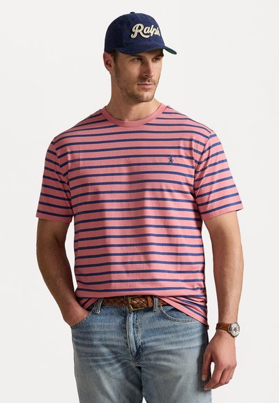 T-shirt rayé rose et marine avec un col rond, des manches courtes et un petit logo sur la poitrine gauche, associé à un jean bleu et une ceinture tissée marron.