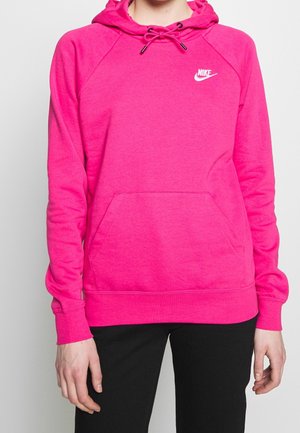 Osoba nosząca jaskrawy różowy kaptur Nike z przednią kieszenią i ściągaczem w kapturze, połączony z czarnymi spodniami, na jednolitym jasnym tle.