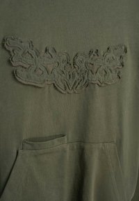 Sweatshirt w kolorze oliwkowym z teksturowanym haftem na klatce piersiowej oraz kieszenią z przodu. Miękka tkanina z lekko wytartym wyglądem.