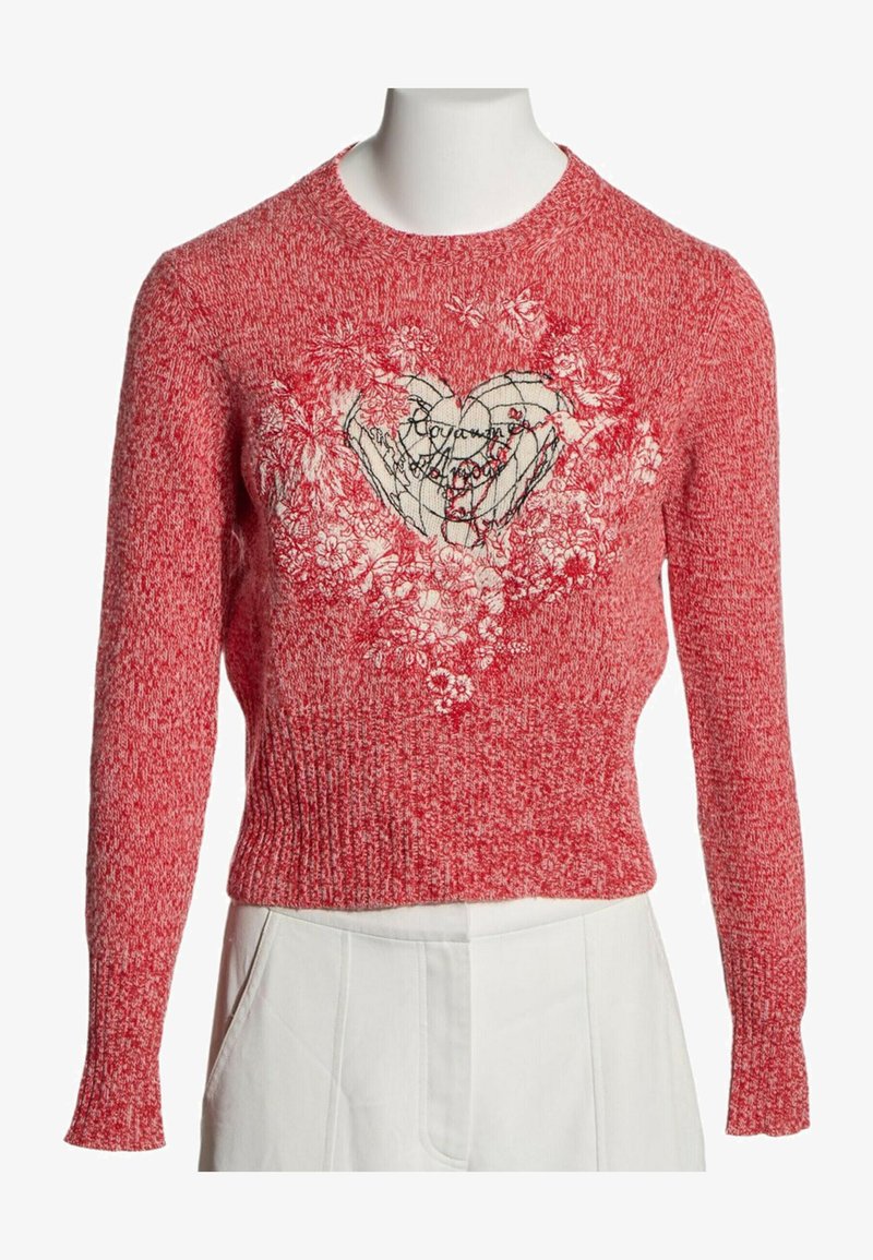 Pull en maille rouge avec un motif cœur blanc entouré de motifs floraux, associé à un pantalon blanc sur un mannequin.