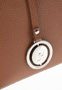 Sac en cuir texturé brun avec un accent en métal argenté en forme de cercle affichant un logo. Dispose d'une bandoulière fermée par un bouton argenté.