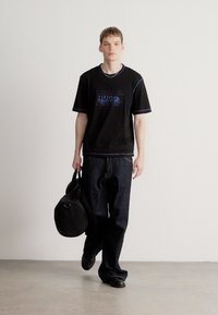 T-shirt preta de mangas curtas com detalhes azuis e logo HUGO, combinada com calças largas de ganga escura. Modelo carrega uma mala redonda preta.