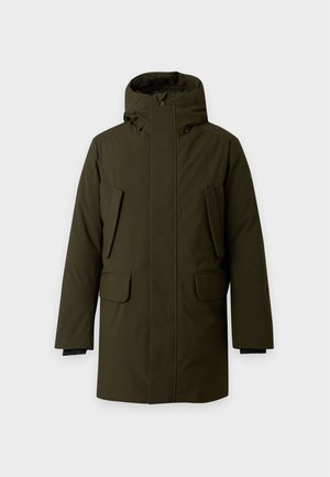Homme debout portant un manteau à capuche vert foncé, une chemise blanche, un pantalon large noir et des chaussures noires sur un fond clair uni.