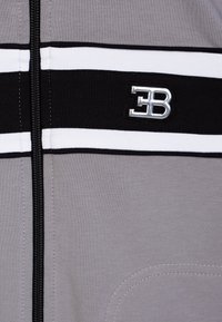 Giacca grigia con zip caratterizzata da righe orizzontali bianche e nere, con un accento logo argentato e una texture morbida. La zip si estende sulla parte anteriore.