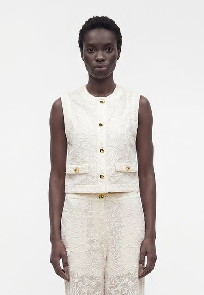 Donna che indossa un gilet bianco senza maniche ricamato con bottoni dorati e pantaloni coordinati con motivo in pizzo, in piedi davanti a uno sfondo uniforme.