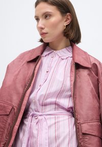 Γυναίκα που φορά χρυσούς κρίκους στα αυτιά, ένα ροζ ριγέ φόρεμα με κουμπιά και δέσιμο στη μέση, και ένα φαρδύ σακάκι σε χρώμα ξεθωριασμένο ρόδι, κοιτάζει προς τα αριστερά.