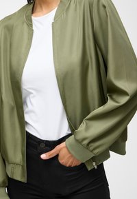 Olivovo zelená bomberka s hladkou, lesklou textúrou, so zipsom, rebrovanými manžetami a okrúhlym výstrihom, v kombinácii s bielou košeľou a čiernymi nohavicami.