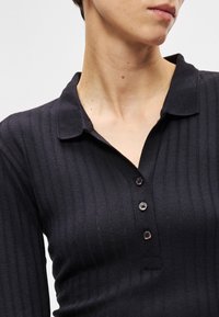 Close-up de uma pessoa a usar uma camisa preta canelada de manga longa com colarinho e três botões, vista do pescoço até à metade do torso.