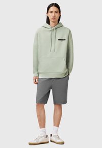 Felpa verde chiaro con cappuccio regolabile, tasca frontale e logo nero. Abbinata a pantaloni corti grigi e sneakers bianche con dettagli beige.