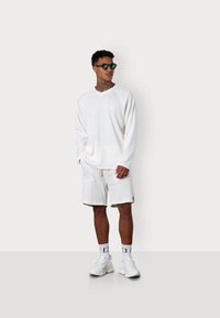 Weißer Pullover mit langen Ärmeln und V-Ausschnitt sowie Raglanärmeln, kombiniert mit passenden weißen strukturierten Shorts. Die Accessoires bestehen aus schwarzen Sonnenbrillen und weißen Turnschuhen.