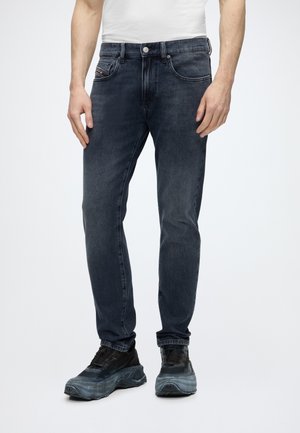 Uomo che indossa jeans slim fit a lavaggio scuro e sneakers nere chunky, in piedi davanti a uno sfondo chiaro e semplice.