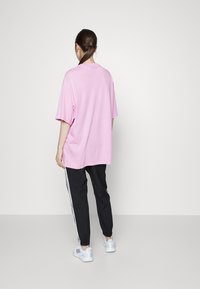 Camiseta de algodón rosa de gran tamaño, pantalones negros entallados con rayas blancas en los costados y zapatillas deportivas blancas, vistos desde atrás contra un fondo gris.