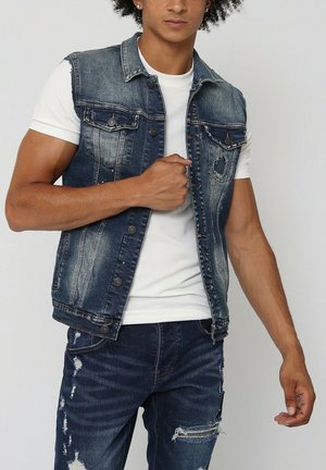 Gilet en jean avec une finition usée, de couleur bleu foncé et boutons-pression. Porté sur une chemise blanche à manches courtes avec une surface texturée.