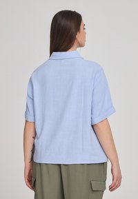 Camicia azzurra a maniche corte con colletto, tessuto testurizzato e maniche arrotolate, abbinata a pantaloni cargo verde oliva. Vista posteriore.