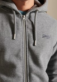 Grijze hoodie met rits, gemaakt van fleece materiaal met een zachte textuur. Voorzien van een capuchon met trekkoord, een zilverkleurige rits en een klein blauw logo.
