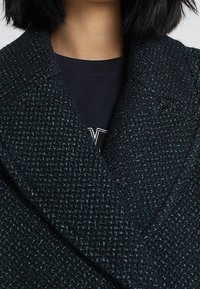 Abrigo de textura verde oscuro con un gran cuello de muesca y patrón de tela tejida. Capa interior visible con una camisa negra debajo.