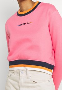 Růžová krátká mikina s modro-oranžovým proužkovaným patentem na límci a spodním lemu. Na přední straně je vyšitý nápis "TOMMY JEANS".