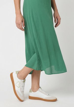 Personne portant une jupe midi verte et des baskets blanches avec des touches vert clair et des semelles en gomme, debout devant un fond blanc uni.