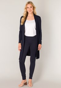 Marineblauwe lange cardigan van zachte stof, gedragen over een wit topje en bijpassende marineblauwe leggings, gecombineerd met beige sandalen met bandjes. Minimalistisch ontwerp.