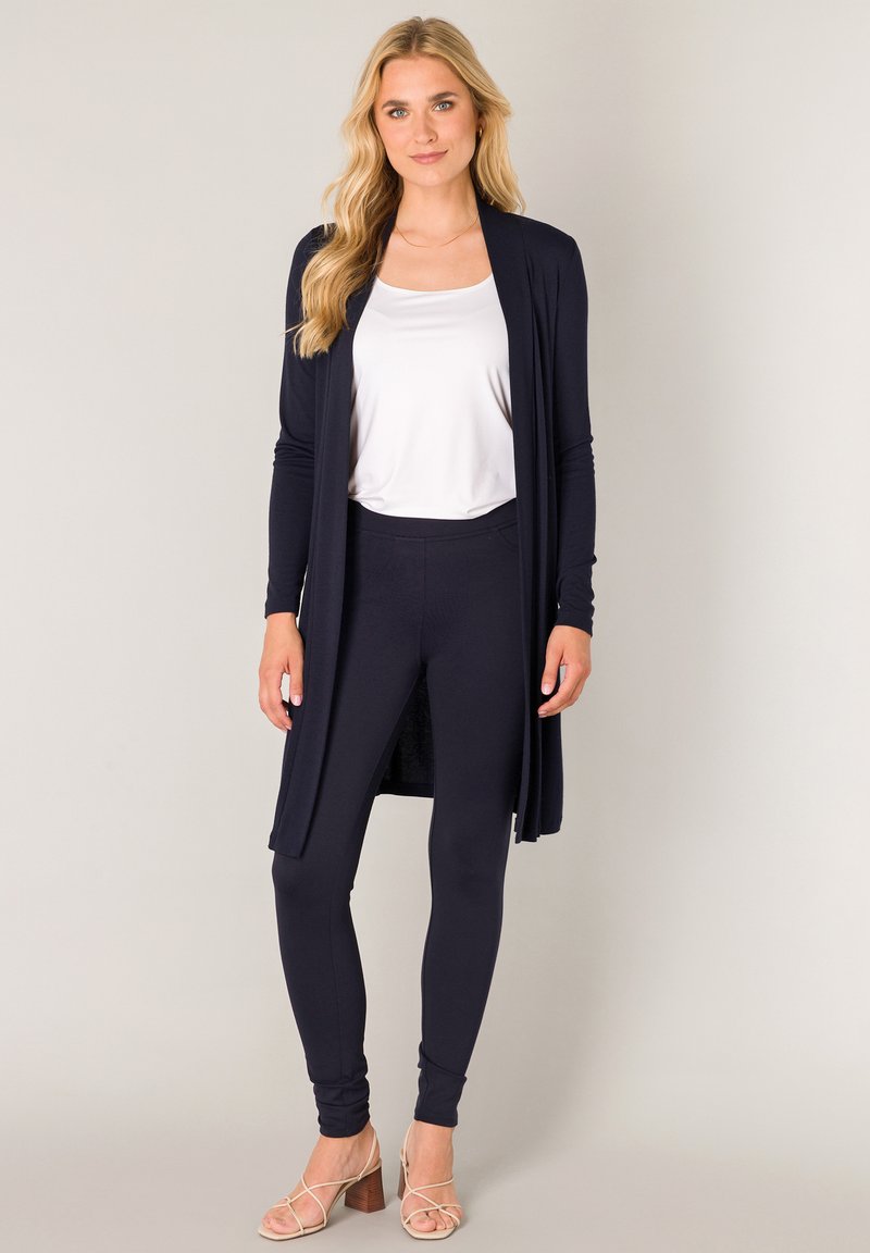 Marineblauwe lange cardigan van zachte stof, gedragen over een wit topje en bijpassende marineblauwe leggings, gecombineerd met beige sandalen met bandjes. Minimalistisch ontwerp.