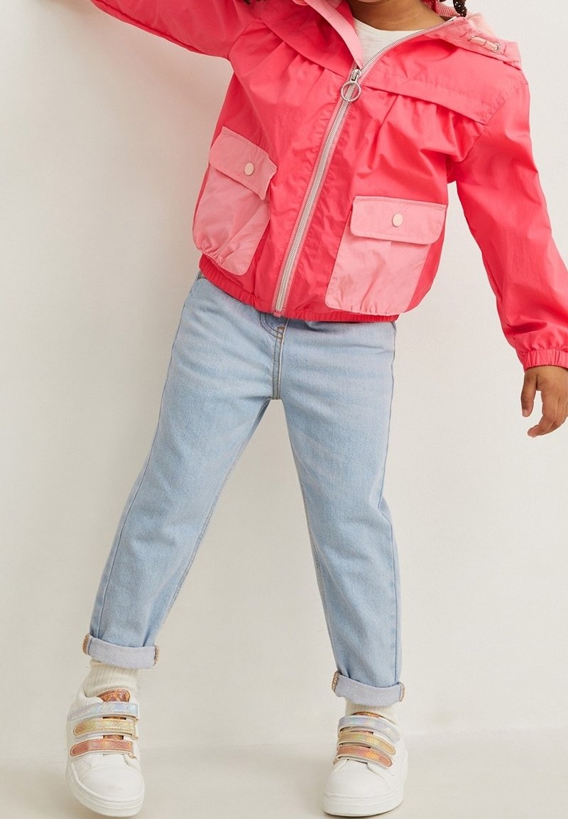 Bambino che indossa un giaccone rosa brillante con tasche a pattina, jeans azzurri arrotolati, calzini bianchi e sneaker bianche con cinturini iridescenti.
