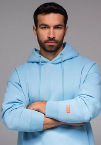 Lichtblauwe hoodie van zachte stof, voorzien van een capuchon met koord, ribgebreide boorden en een klein oranje merkje op de mouw.