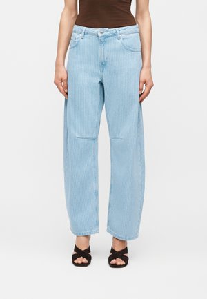Kvinde iført lyseblå jeans med brede ben og fine striber, kombineret med sorte peep-toe hæle med krydsede stropper og en brun ærmeløs top.