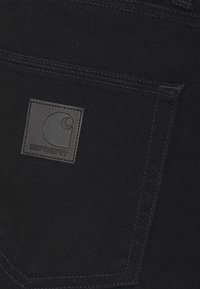 La tela de mezclilla negra presenta una etiqueta visible grabada con el logo de Carhartt, ubicada en el bolsillo trasero de la prenda.